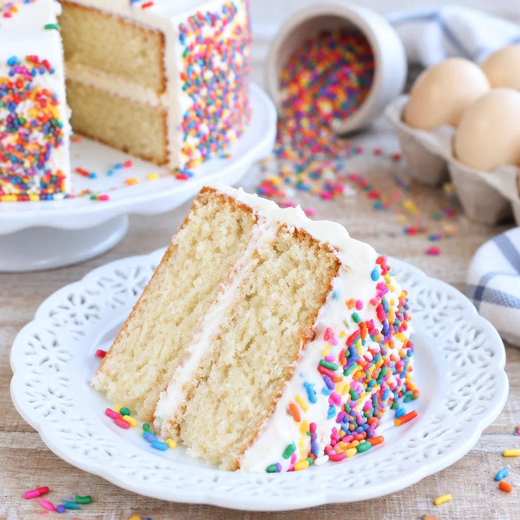 A slice of moist vanilla cake topped with rich vanilla buttercream and sprinkles.
